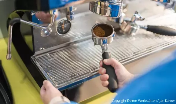Eine Person führt den gefüllten Siebträger zur vorbereiteten Kaffeemaschine (Copyright: Delme-Werkstätten/Jan Karow)